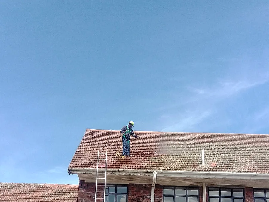 A Roof Direct worker cleaning a weathered clay tiled roof on a red-brick building, before painting