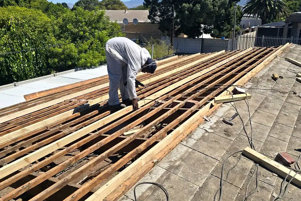 A worker in a protective suit fixing new timber battens as an old asbestos cement roof comes off