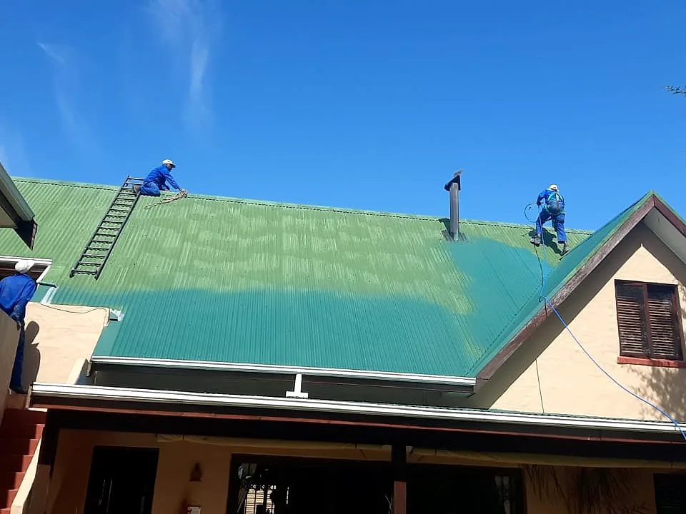 Two Roof Direct painters on ladders applying green paint to a corrugated metal house roof